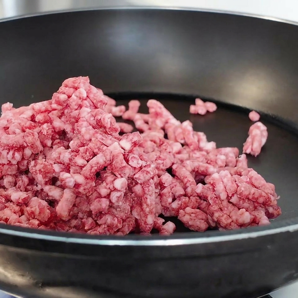 Pink raw minced beef being broken up and stir-fried in a black non-stick pan.