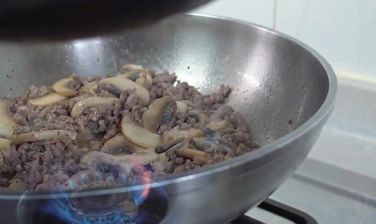 A stainless steel wok containing a mixture of browned ground beef and sautéed mushroom slices being tossed over a gas flame.