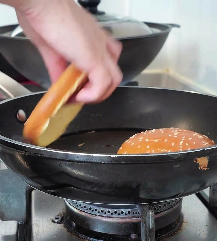 A hand placing a sesame seed burger bun half into a black skillet next to another toasting bun.