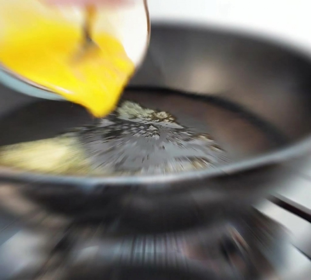 Beaten yellow egg liquid being poured from a bowl into a hot pan with melted butter.