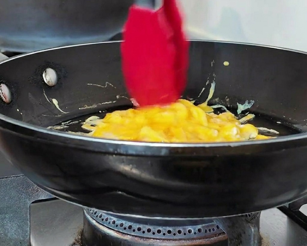 Red tongs gathering soft scrambled eggs into a circular shape in a black pan.