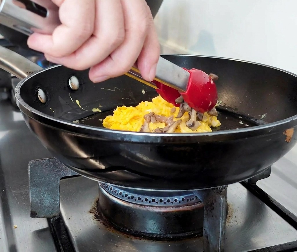 Cooked minced beef and sliced mushrooms being piled onto a bed of soft scrambled eggs in a pan.