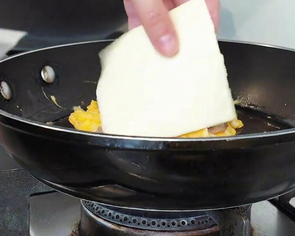 A hand placing a square slice of white cheese onto a mound of beef and eggs in a frying pan.