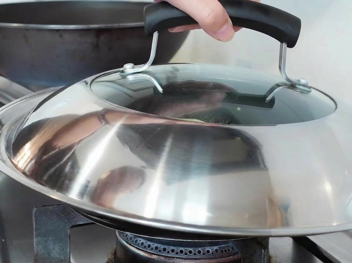 A hand placing a large metal and glass lid over a frying pan on a gas stove.