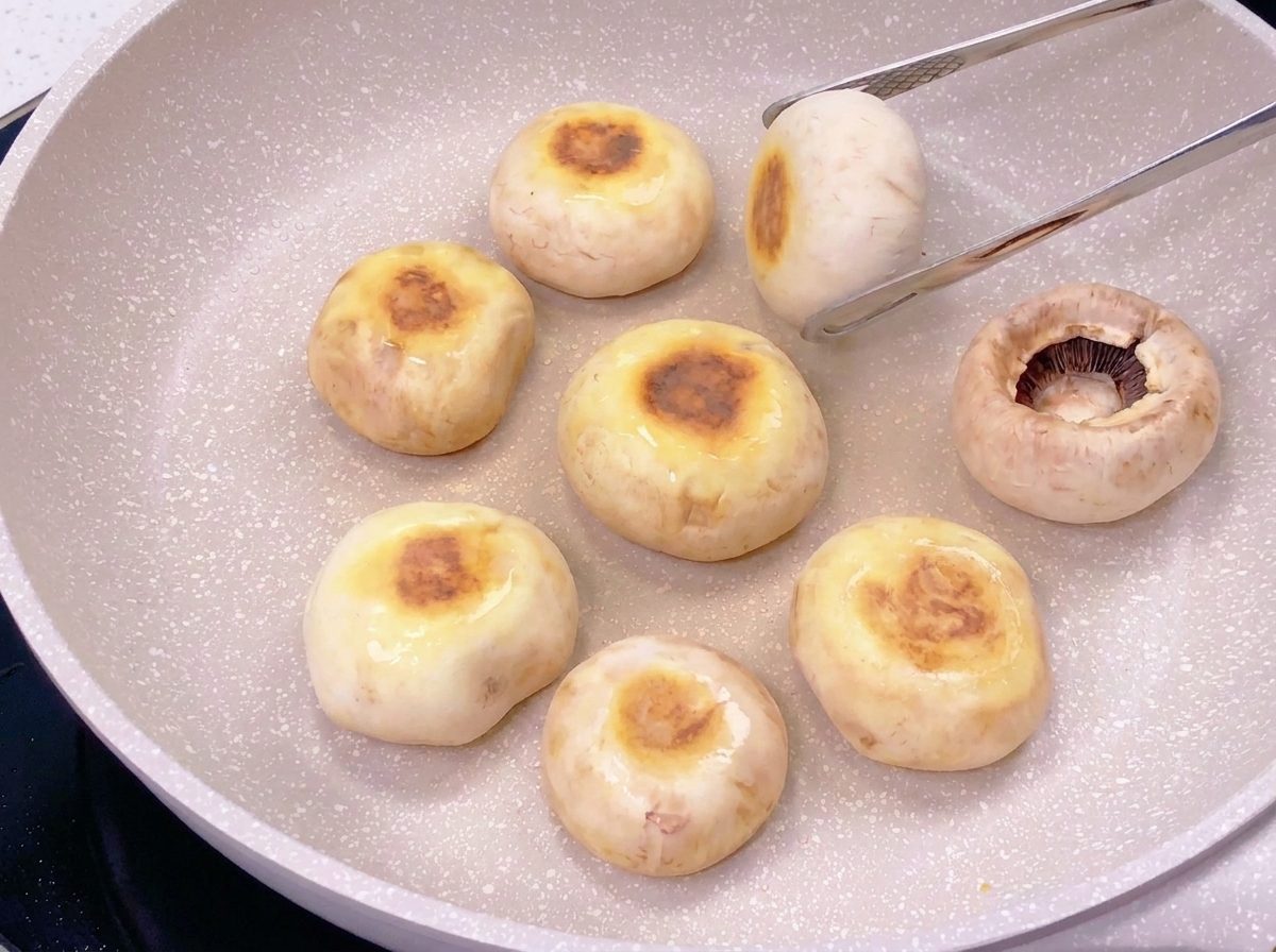 Seven mushroom caps in a frying pan being turned over to reveal circular brown sear marks on their tops.