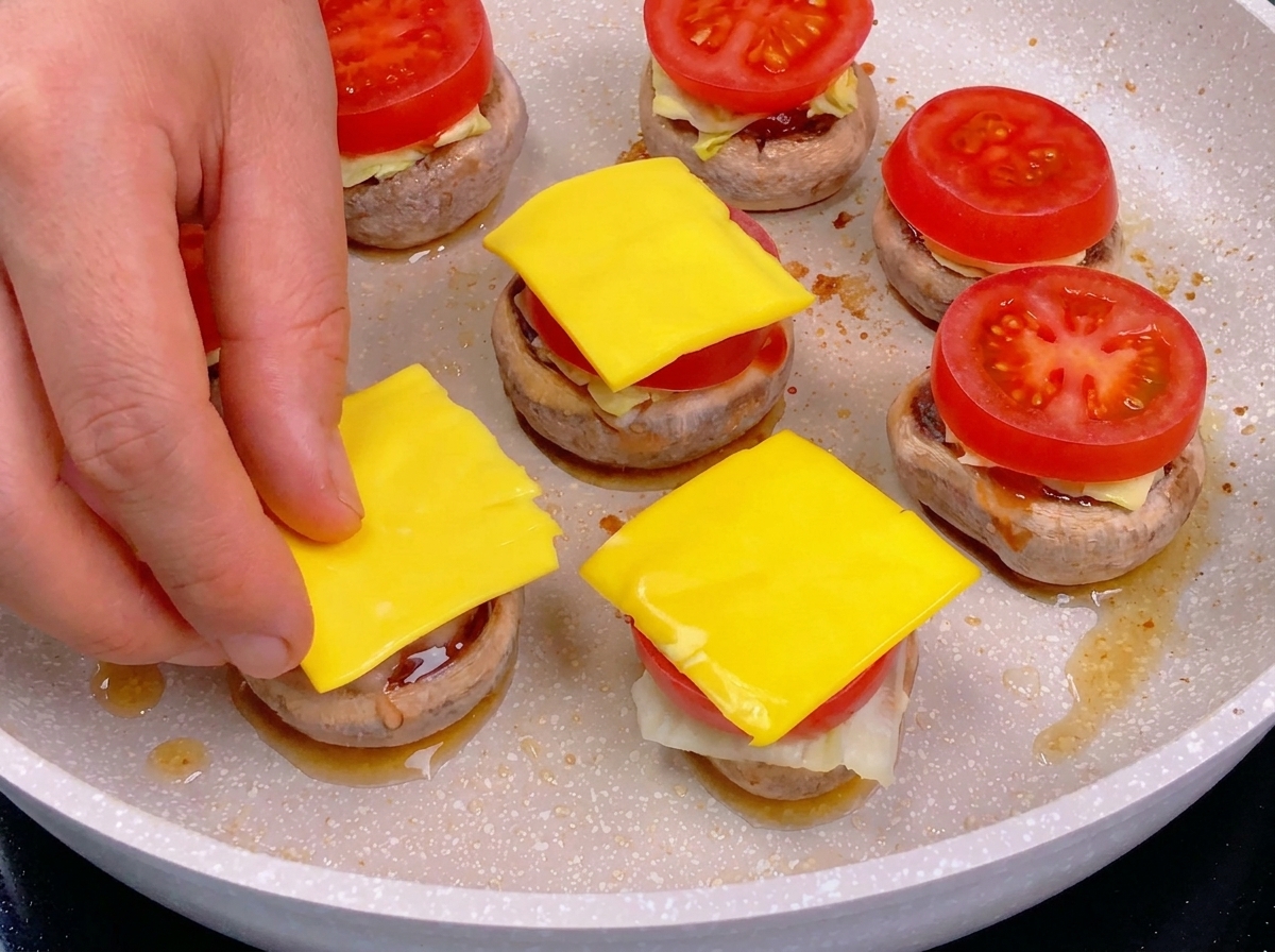 A hand placing a square slice of yellow cheese onto a tomato slice as part of a layered mushroom burger assembly.