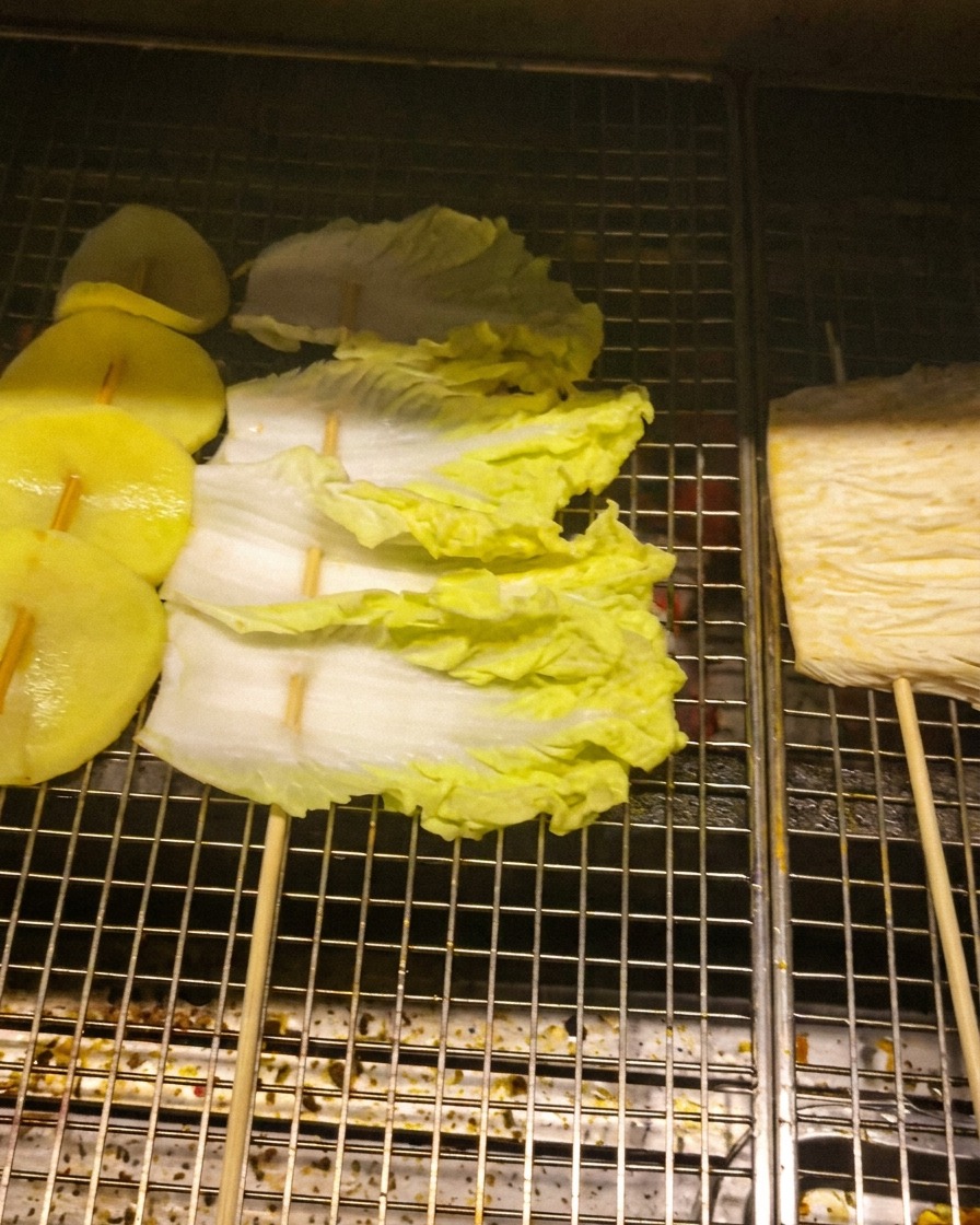 Slices of potato, cabbage leaves, and enoki mushrooms threaded onto skewers and laid across a metal grill rack.