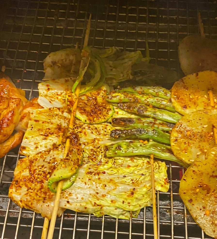 A close-up of grilled chicken, cabbage, and zucchini being brushed with a rich red BBQ sauce and garnished with green onions.