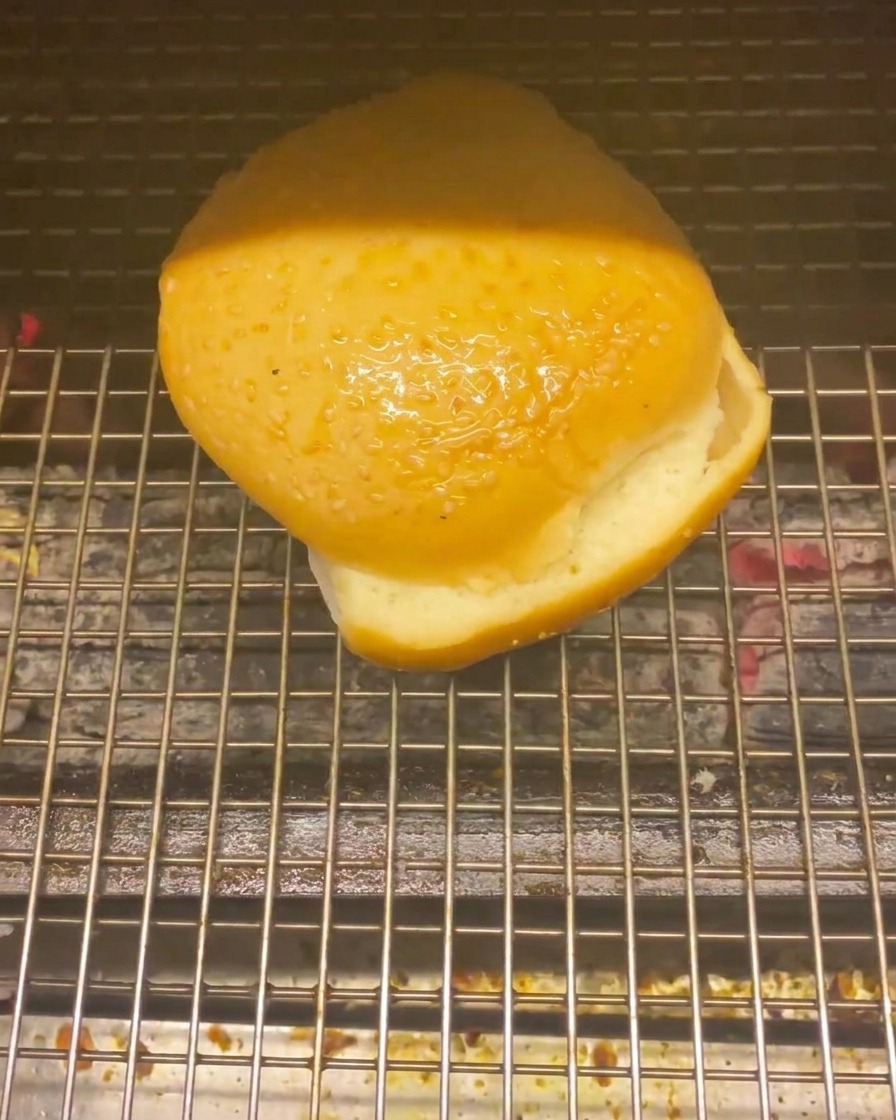 A burger bun placed on a wire grill over hot coals to toast.