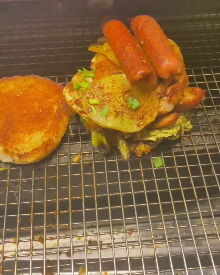 A tall stack of grilled meats and vegetables, including sausages and potato rounds, piled onto a burger bun on a wire rack.