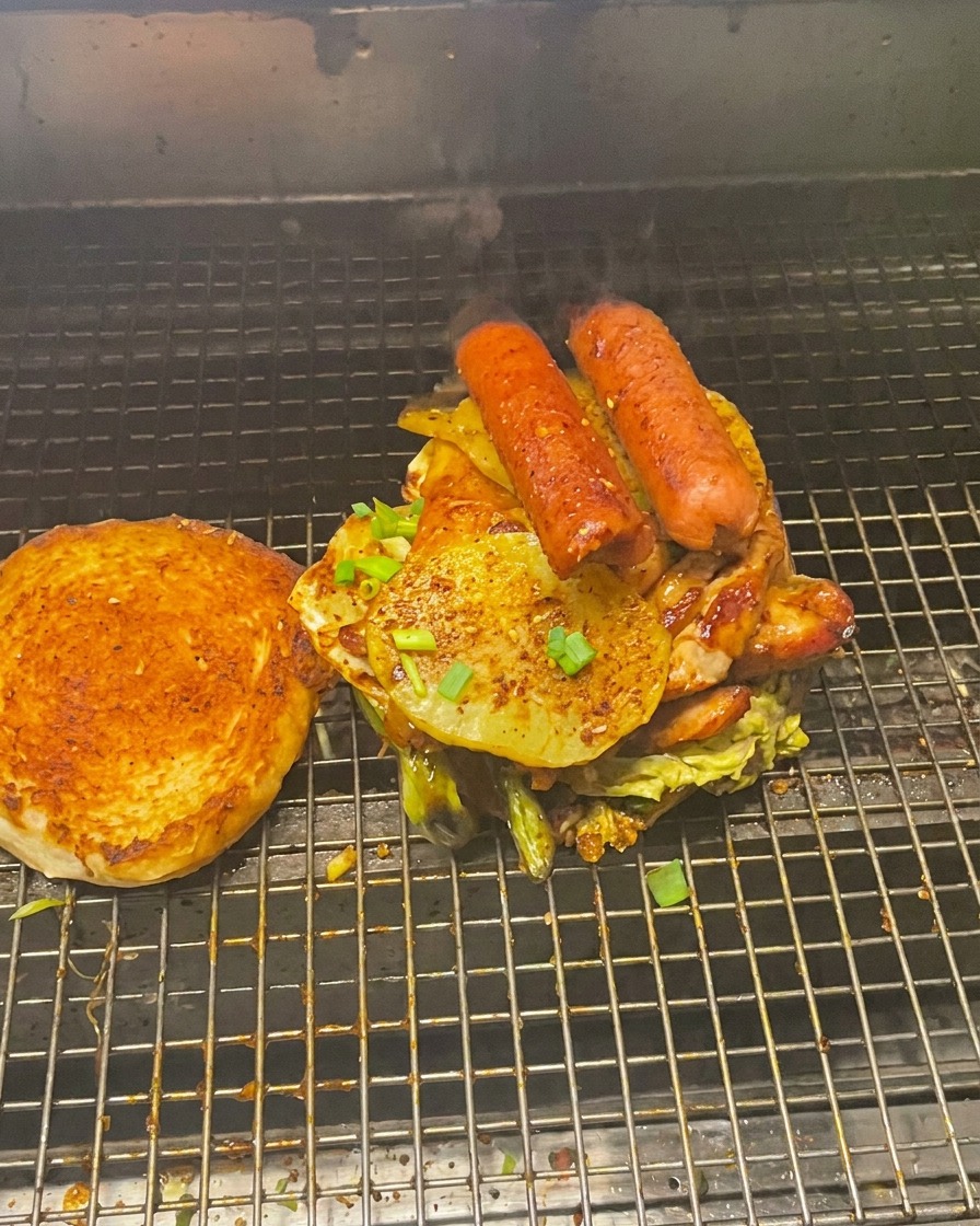 A towering stack of grilled meats and vegetables on a bottom bun, with the toasted top bun resting beside it on the grill.