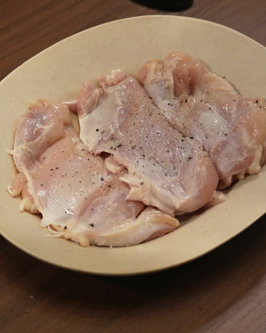 Raw chicken thighs laid out on a beige plate, sprinkled with black pepper and various ground spices.