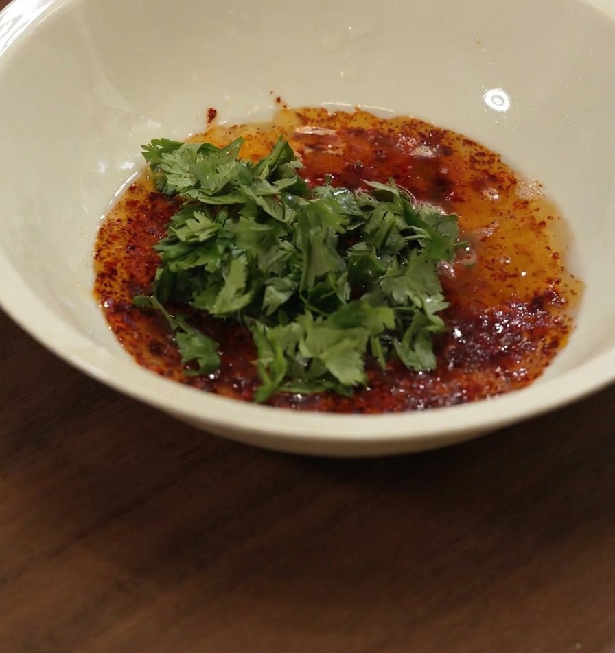 Honey being poured into a white bowl containing a mixture of red oil, spices, and chopped green cilantro.