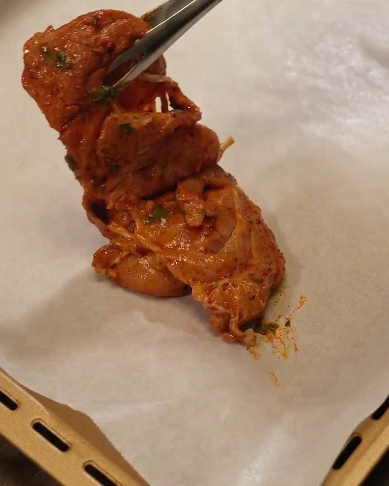 Metal tongs lifting a marinated chicken thigh and placing it onto a baking sheet lined with white parchment paper.