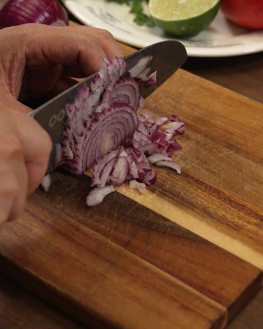 A hand using a large kitchen knife to finely dice a red onion on a sturdy wooden cutting board.