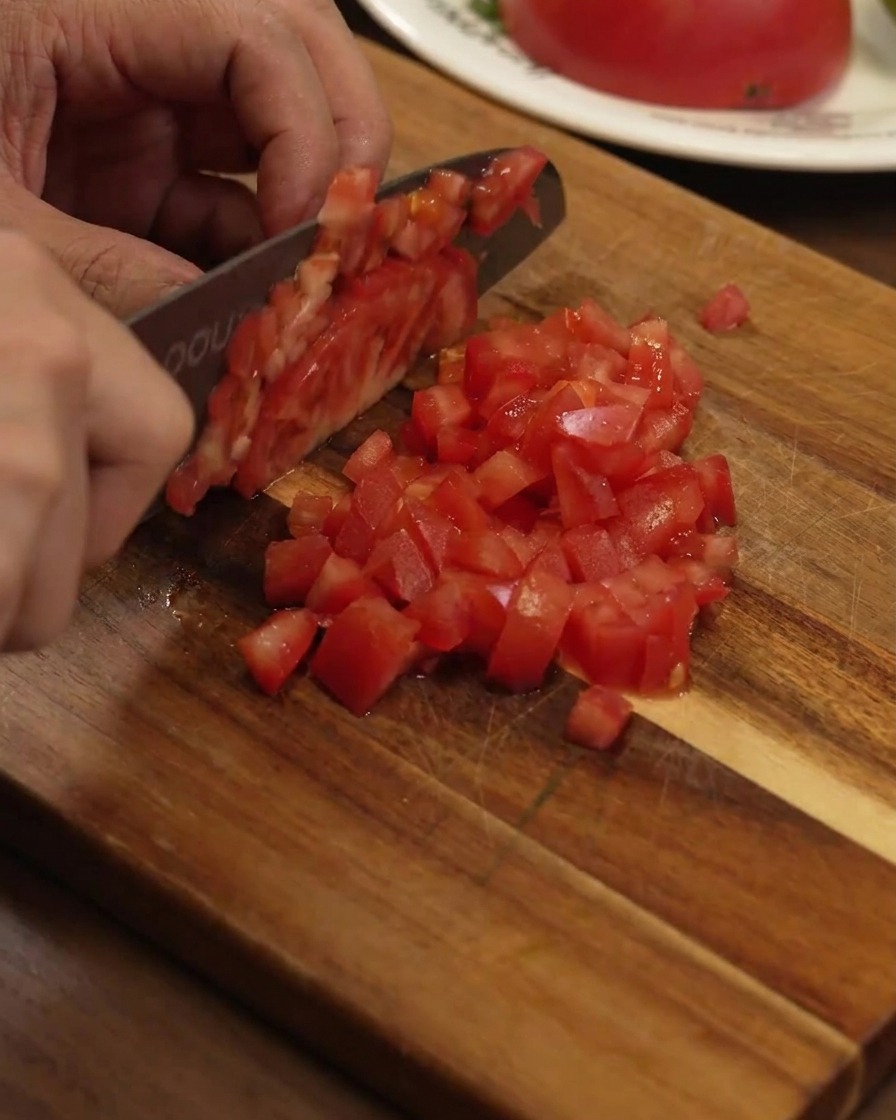 Dicing a ripe red tomato into small even cubes on a wooden board using a chefs knife.