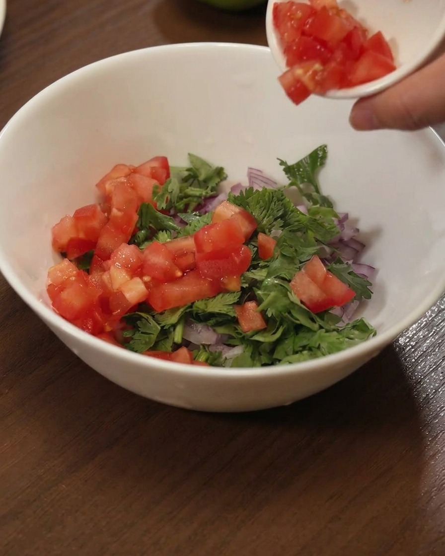 Diced red tomatoes being poured from a small dish into a white bowl filled with green cilantro and purple onions.