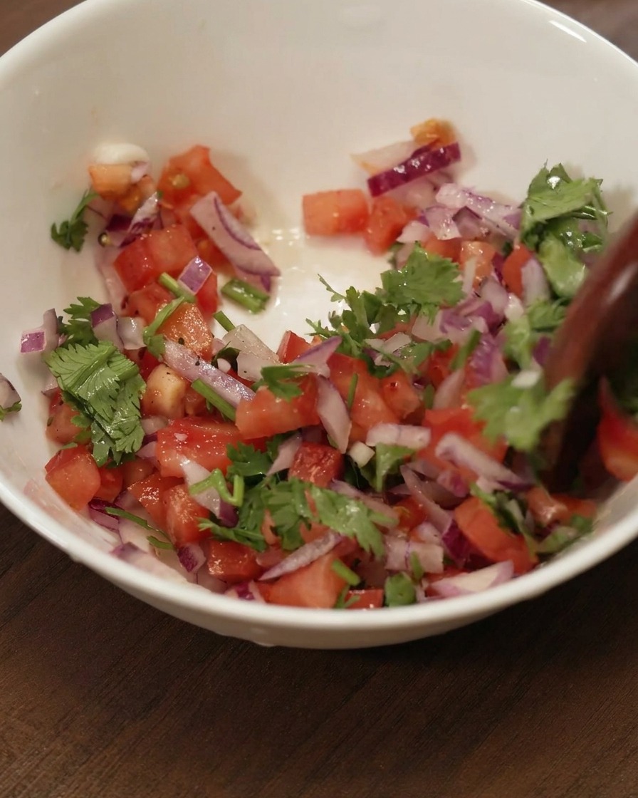 A wooden spoon stirring a colorful mixture of diced tomatoes, red onions, and cilantro in a white ceramic bowl.