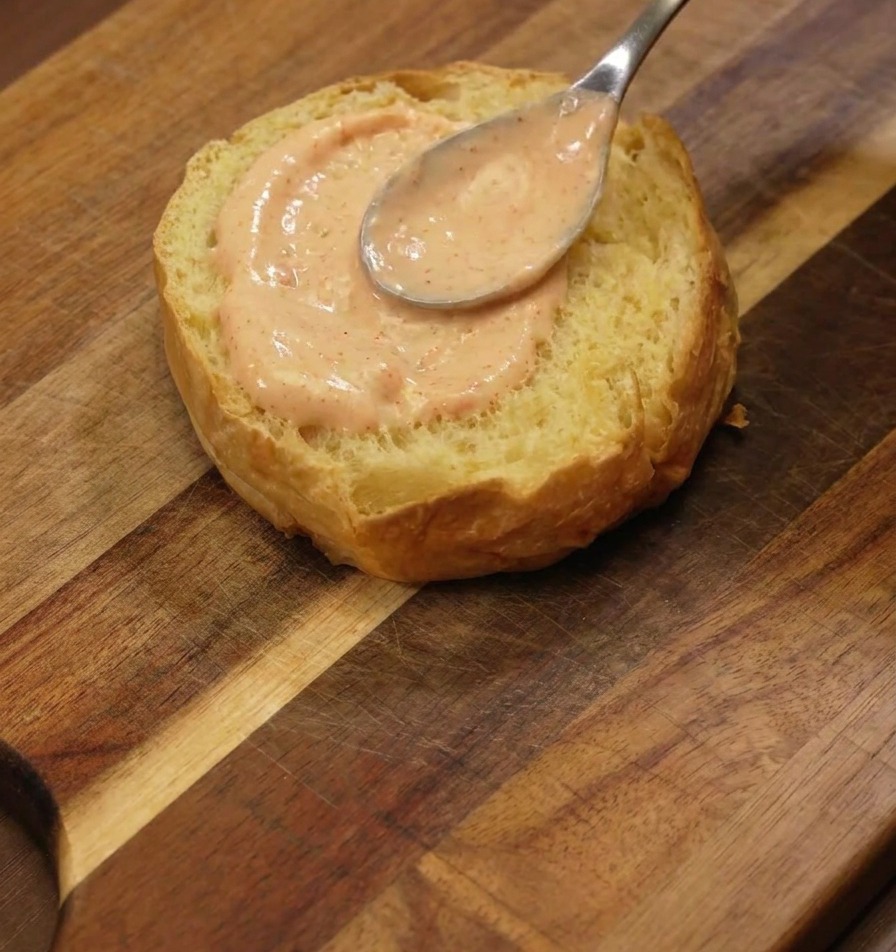 A silver spoon spreading a thick layer of creamy orange burger sauce onto the toasted face of a brioche bun on a wooden cutting board.