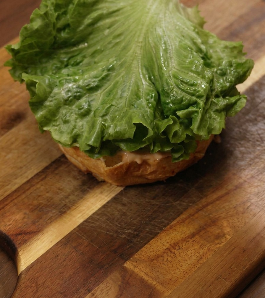 A vibrant green leaf of lettuce resting on top of a sauced burger bun on a wooden board.