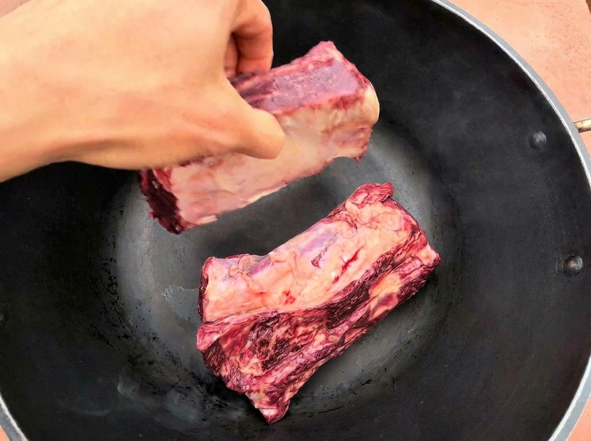 A hand placing two large, raw beef ribs into a hot black pan.