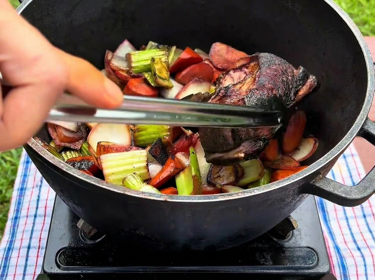 A hand using metal tongs to place a deeply seared beef rib into a black pot filled with sautéed vegetables.