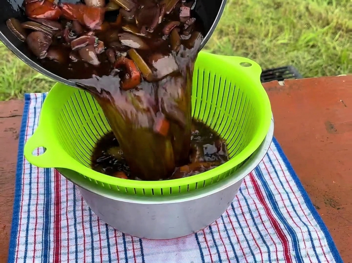 Rich, dark braising liquid along with cooked vegetables being poured from a pot into a bright green strainer sitting over a stainless steel bowl.