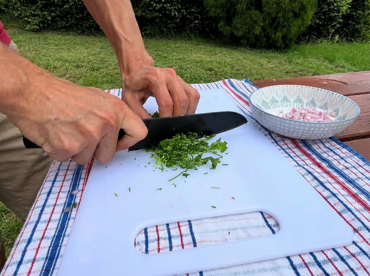 A persons hands using a black knife to finely chop fresh green parsley on a white cutting board next to a small bowl of diced shallots.