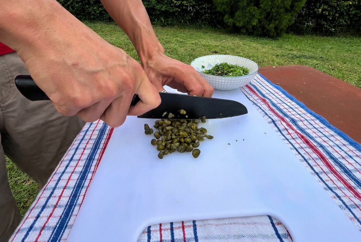 A close-up view of a person using a black chefs knife to finely chop green capers on a white cutting board outdoors.