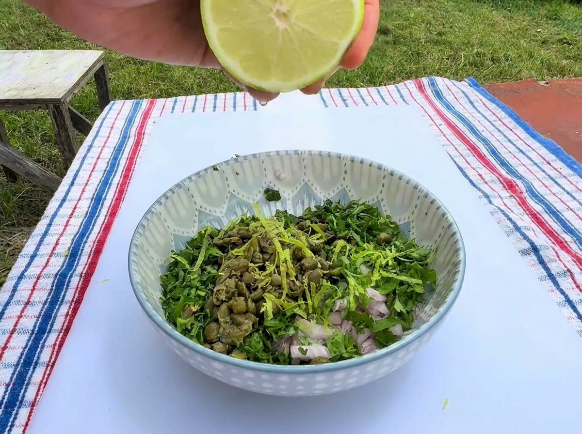 A hand squeezing half of a fresh lime over a patterned ceramic bowl filled with chopped green herbs, capers, minced shallots, and bright lime zest.