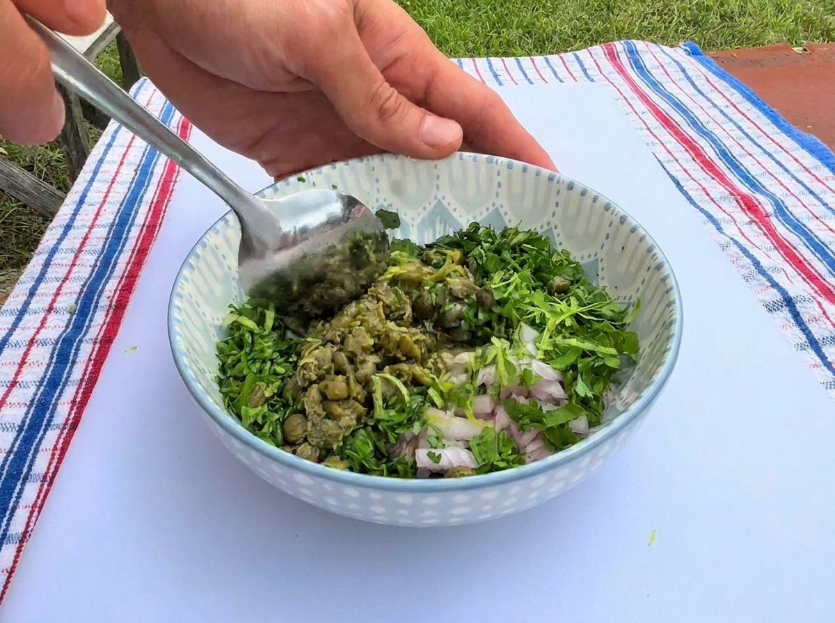Stirring a vibrant mixture of chopped green herbs, capers, and minced shallots with a metal spoon inside a patterned ceramic bowl.
