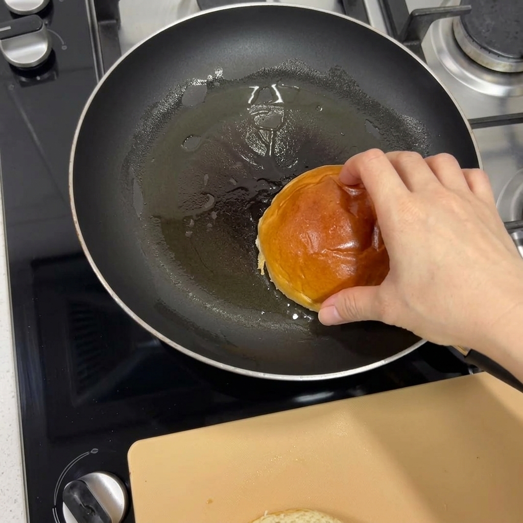 A hand placing a brioche bun into a heated frying pan to toast it until golden brown.
