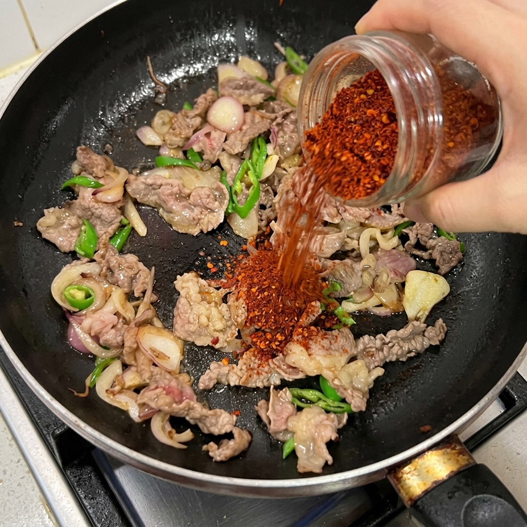 Red chili powder being poured from a glass jar onto sliced beef, onions, and green chilies stir-frying in a black pan.