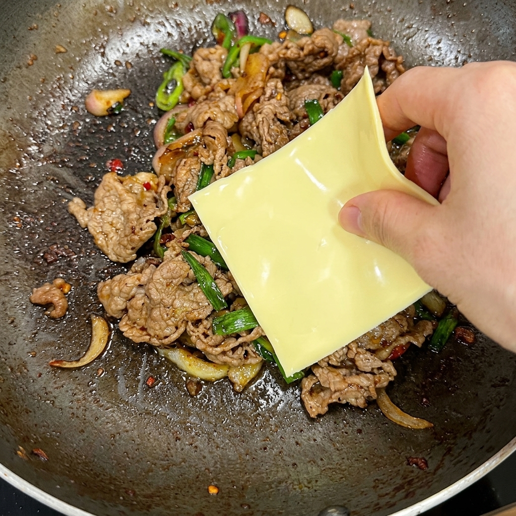 A hand placing a square slice of yellow cheese on top of a hot stir-fried beef and green pepper mixture in a pan.
