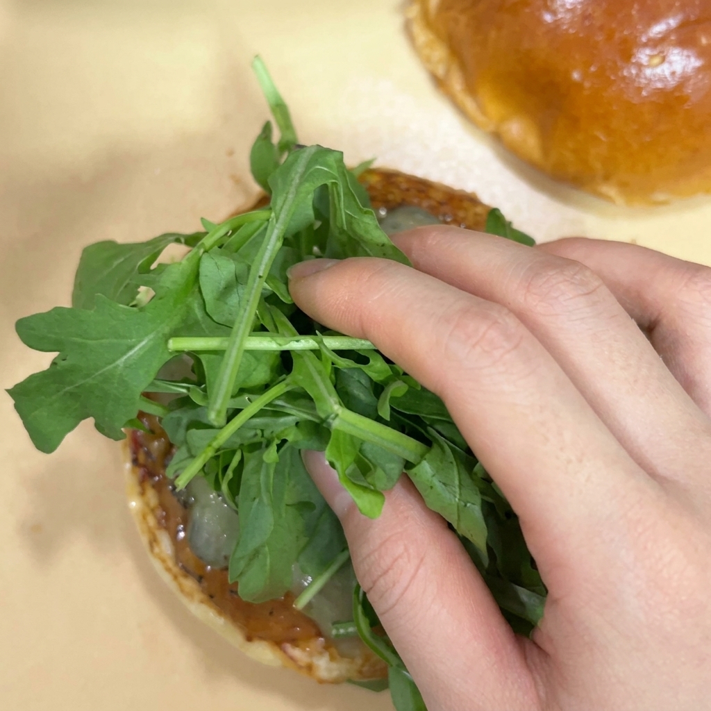 A hand placing a fresh bunch of green arugula leaves over pickles on a toasted burger bun half.