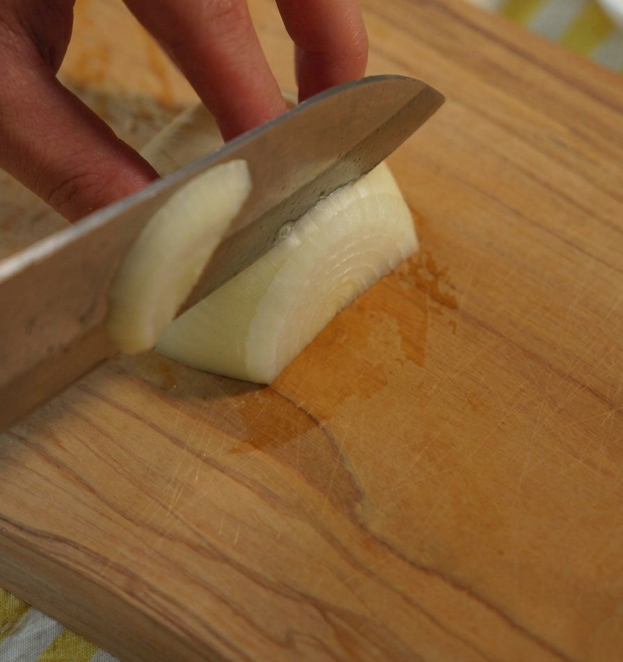 A close up shot of a hand holding a white onion while a sharp chefs knife slices it into uniform pieces on a wooden board.