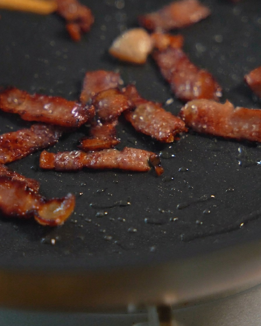 Small pieces of diced bacon sizzling in a black frying pan, showing various stages of browning and crisping.
