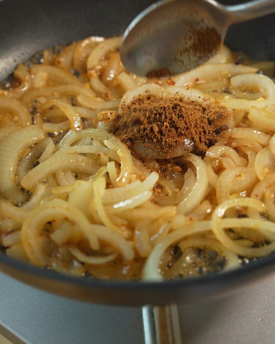 Slices of onion being sautéed in a pan with a spoon adding brown sugar and liquid seasonings for caramelization.