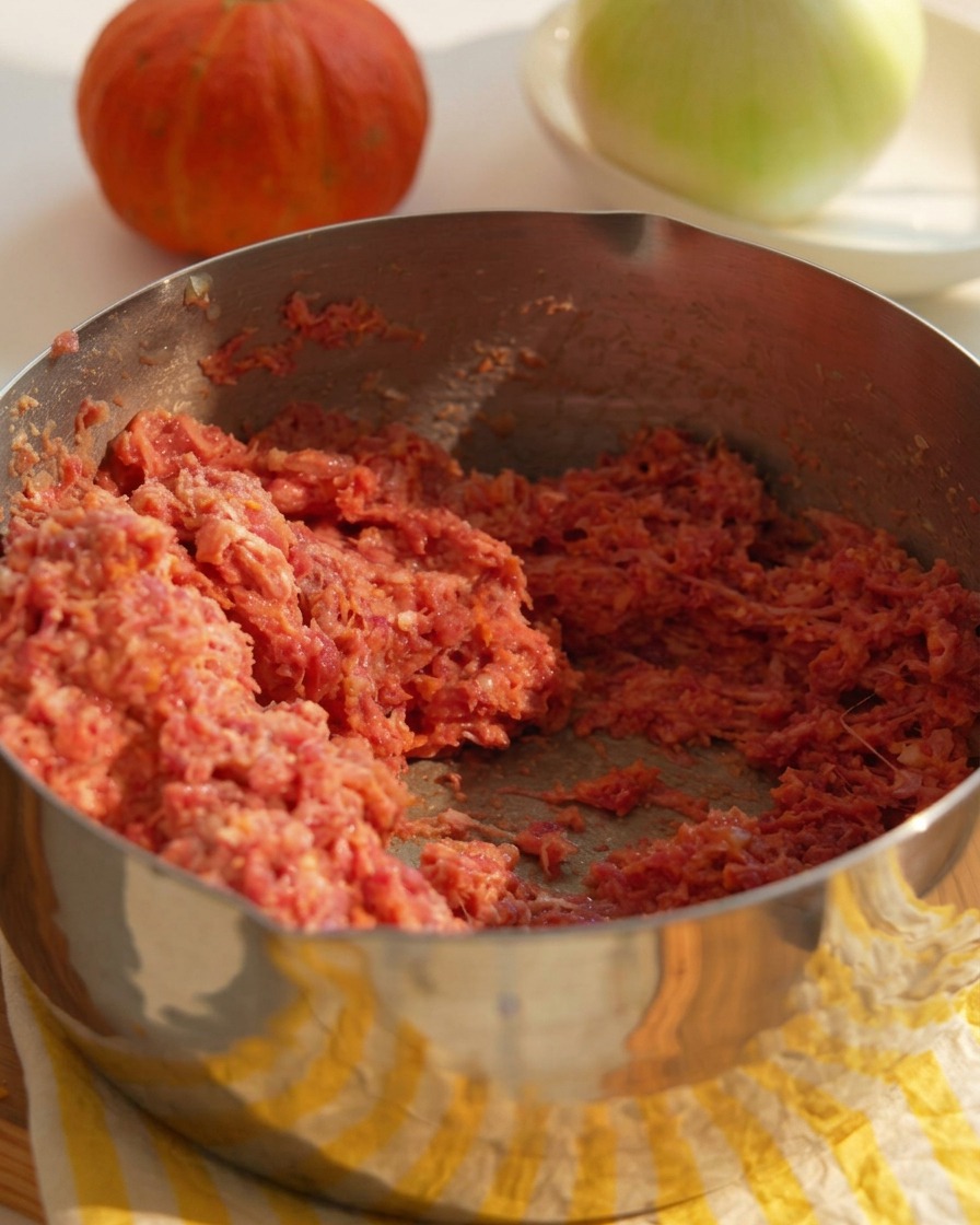 A metal bowl filled with a mixture of raw ground beef, minced onions, and binder ingredients being prepared for burger patties.