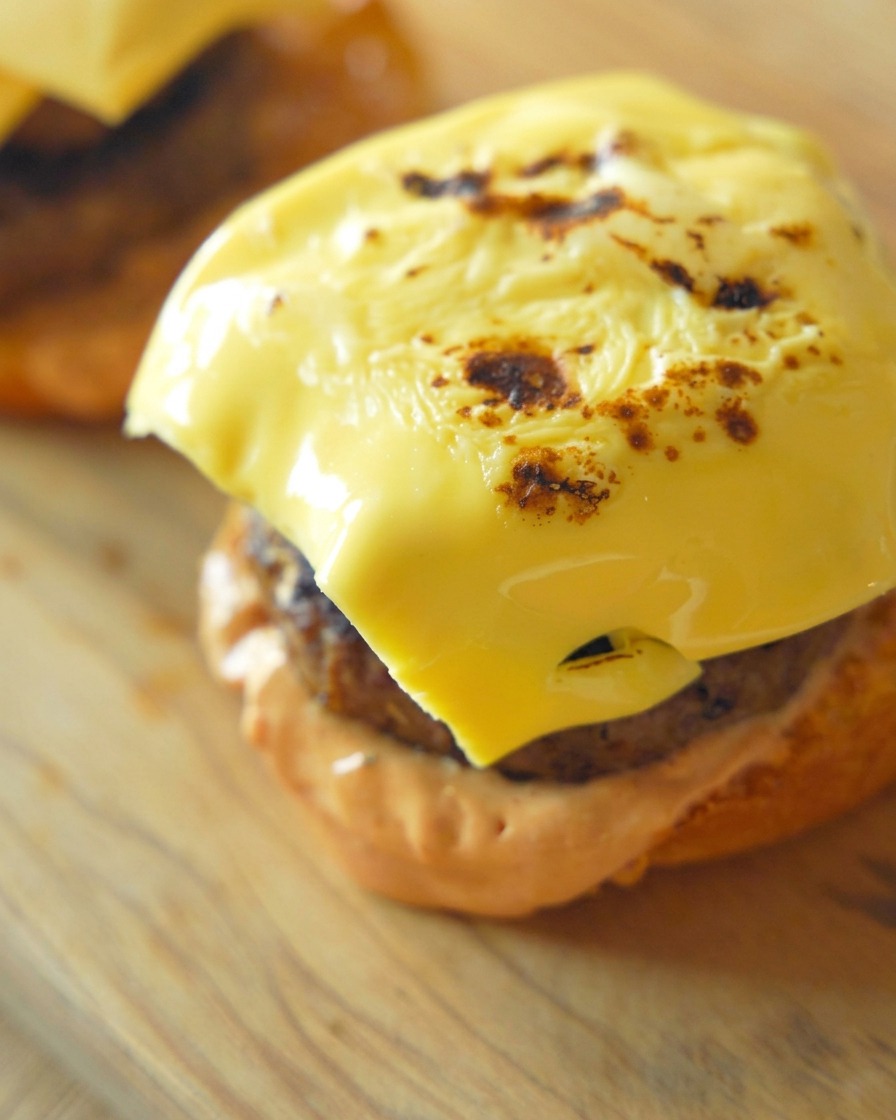 A close up of a perfectly melted and slightly charred yellow cheese slice draped over a thick cooked beef patty on a bun base.