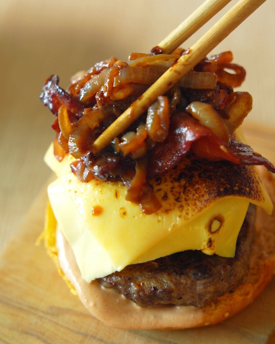 Crispy bacon strips and sweet caramelized onions being carefully placed onto a cheese-topped burger patty using wooden chopsticks.