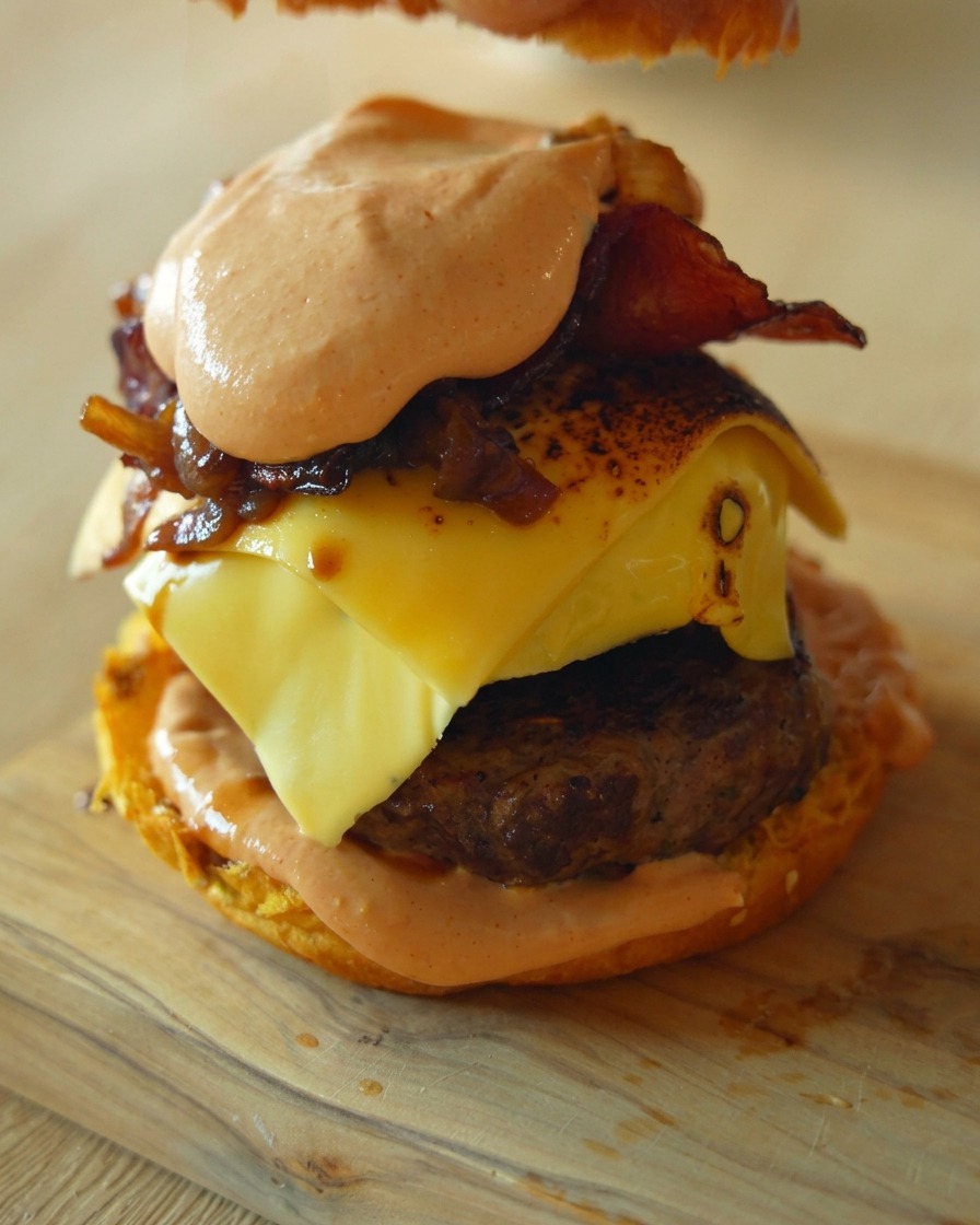 A close-up shot showing a hand placing the final top bun onto a tall, gourmet burger stacked with a beef patty, melted cheese, and a generous portion of caramelized onions and sauce.