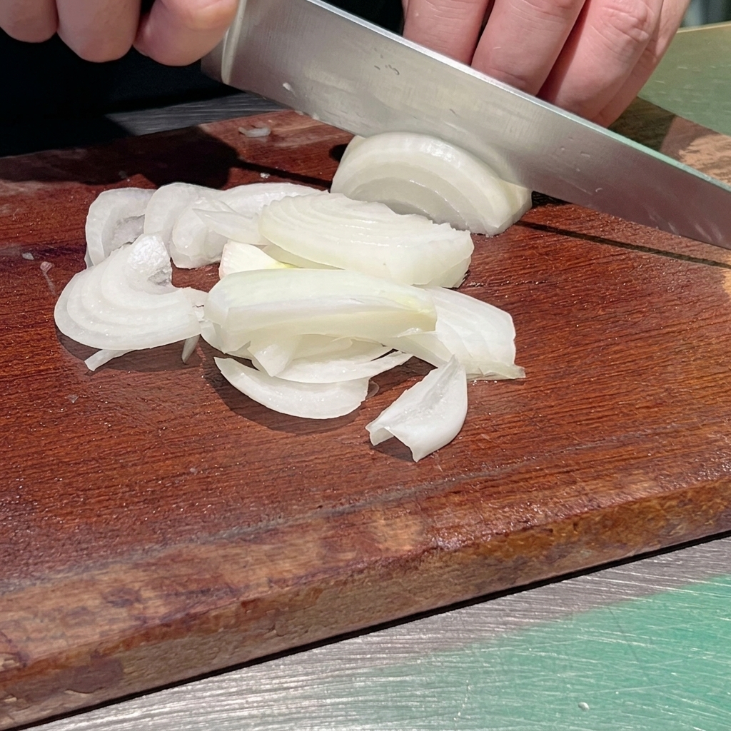 A close-up shot of a chef using a sharp kitchen knife to slice a white onion into thin strips on a dark wooden cutting board.