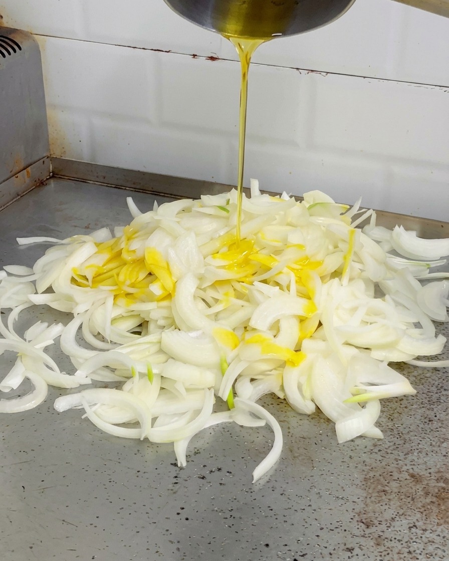 Melted yellow butter being poured from a silver pot onto a pile of white onions on a flat-top grill.