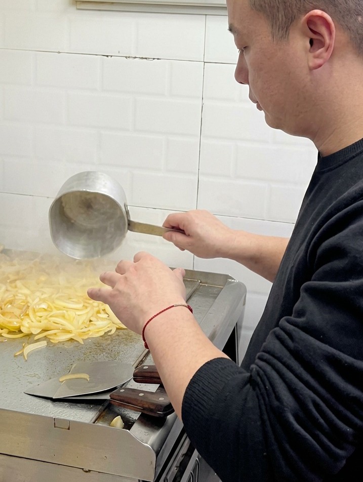 A chef pouring water from a small metal ladle onto a pile of softening onions on a commercial flat-top grill.