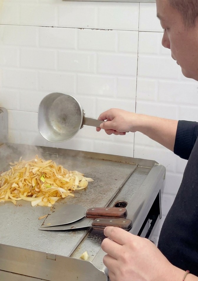 A chef adding more water to a pile of light brown, partially caramelized onions on a grill surface.