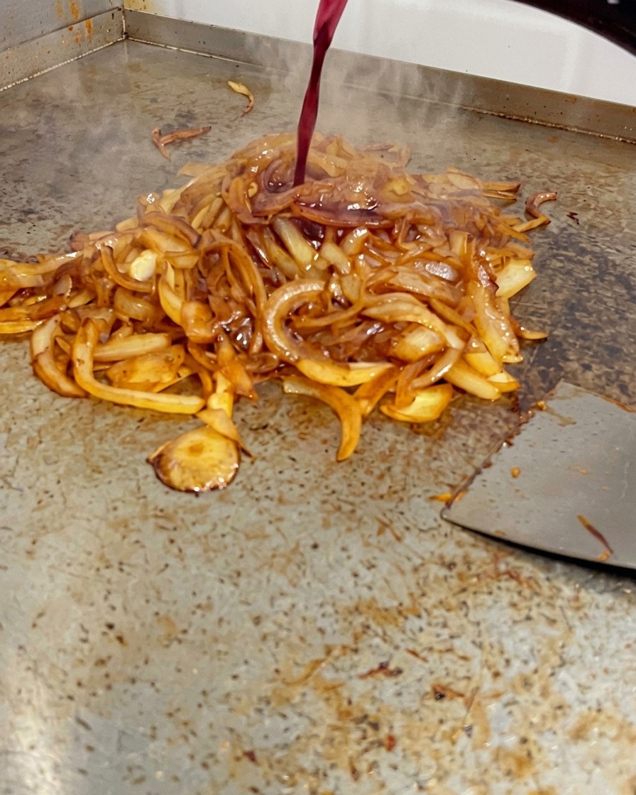 Red wine being poured from a bottle onto a pile of deeply browned onions on a flat-top grill.