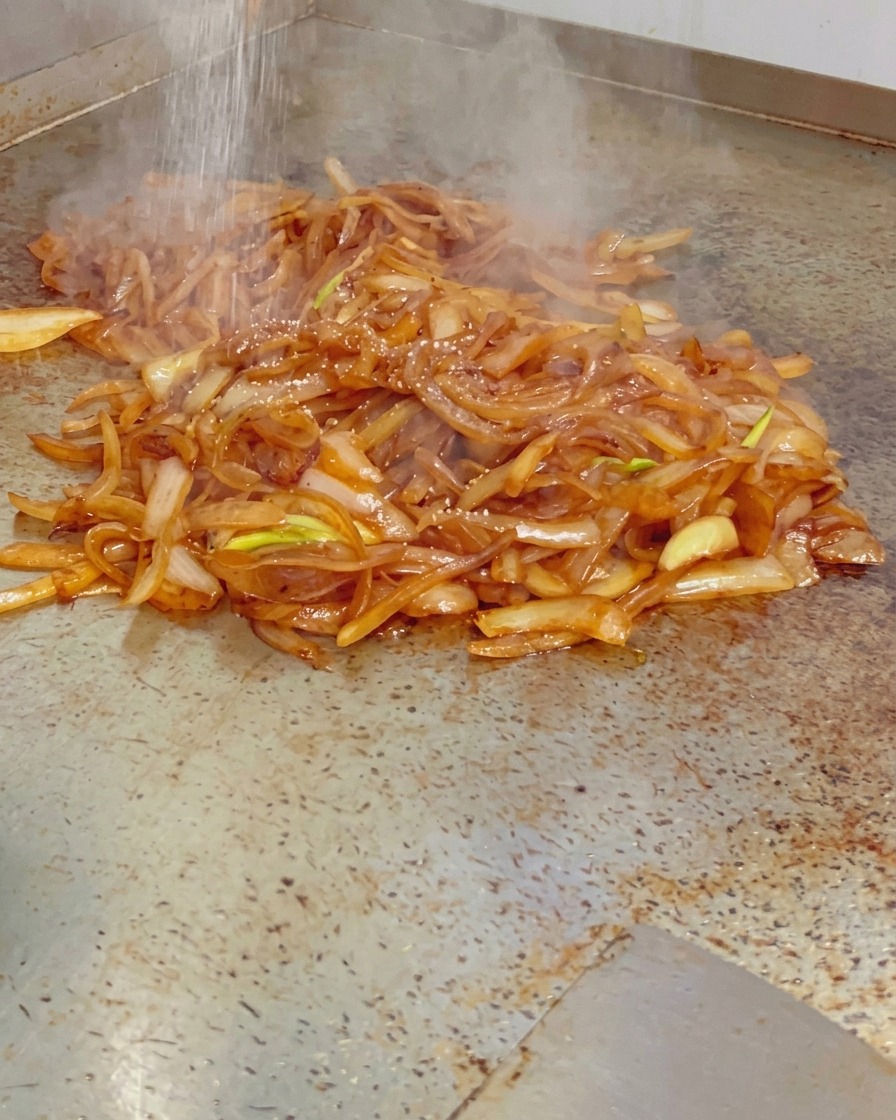 Fine salt being sprinkled over a pile of steaming, glossy caramelized onions on a hot flat-top grill.