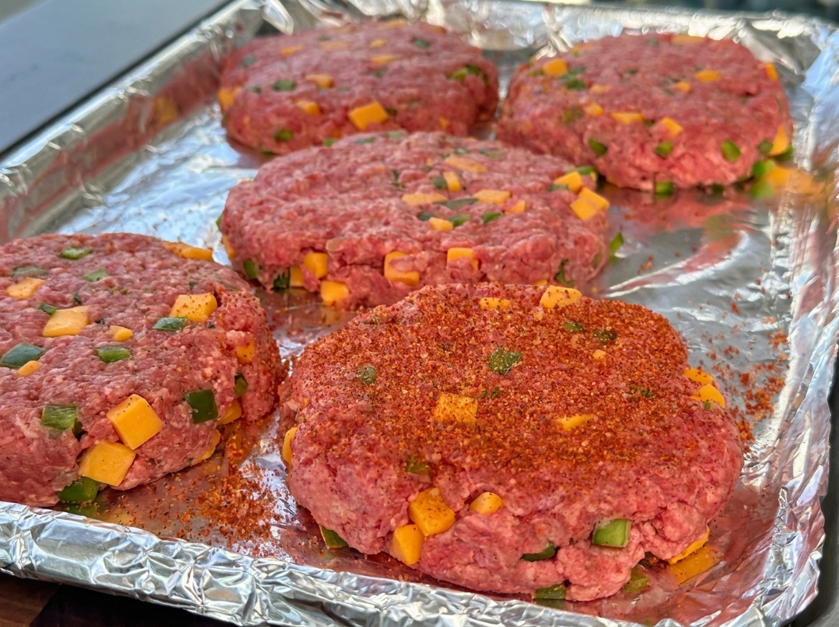Formed raw beef burger patties with visible cheese cubes and green peppers, seasoned with red BBQ powder on a foil tray.