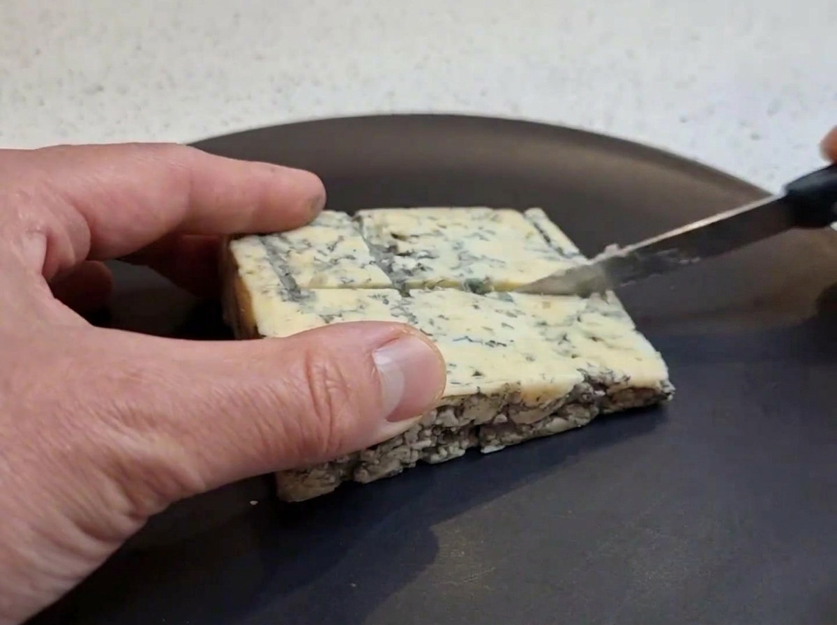 A hand using a small knife to cut a square block of blue cheese into smaller chunks on a dark plate.
